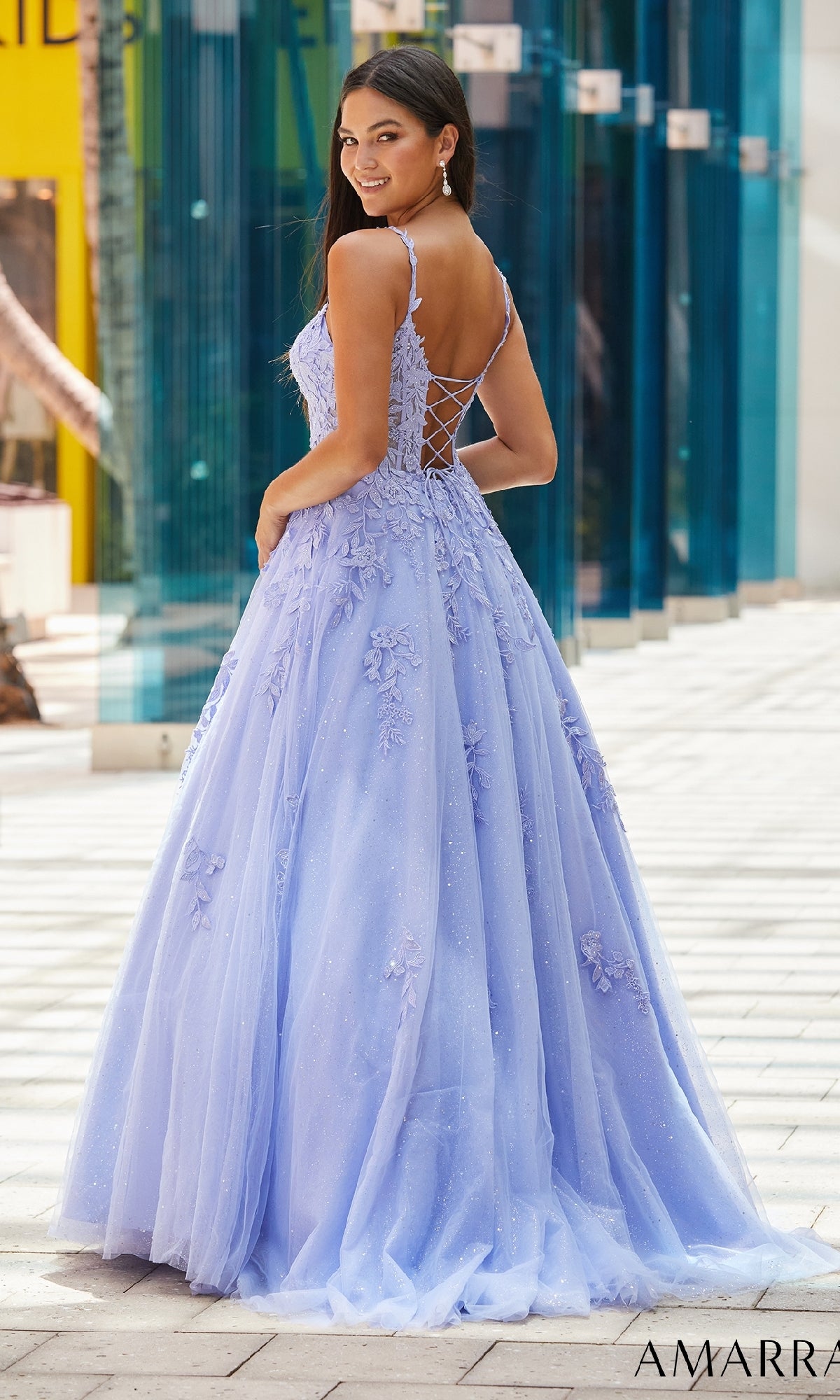A woman wearing the Amarra 88590 Floral Lace Prom Ball Gown, a light purple floor-length dress with a lace-up back, stands outdoors and looks over her shoulder.